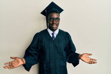 Mezuniyet şapkası ve tören cübbesi giyen yakışıklı siyah adam gülümseyerek avuçlarını açıyor, sunuyor ve reklam karşılaştırması yapıyor ve dengeyi sağlıyor. 