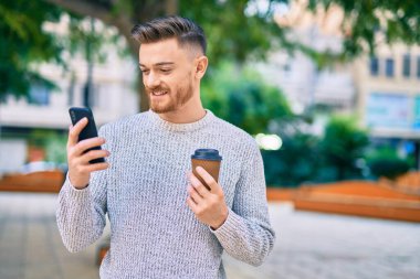 Genç beyaz adam akıllı telefon kullanıyor. Parkta kahve içiyor..