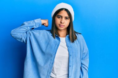 Şirin yün şapkalı genç Latin kadın güçlü kol kası gösteren, kendinden emin ve güçle gurur duyan biri. 