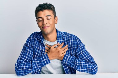 Genç, yakışıklı, Afro-Amerikan bir adam, gündelik giysiler içinde masada oturmuş, elleri göğsünde, gözleri kapalı ve yüzünde minnet dolu bir jest ile gülümsüyor. sağlık kavramı. 