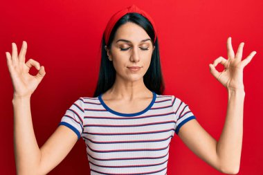 Gündelik kıyafetler giyen, gözleri kapalı gülümseyen genç İspanyol kadın parmaklarıyla meditasyon hareketi yapıyor. yoga konsepti. 