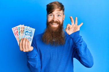 Genç İrlandalı kızıl saçlı adam Yuan Çin banknotlarını tutuyor tamam işareti yapıyor parmaklarıyla, gülümseyerek mükemmel bir sembol oluşturuyor 