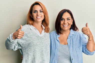 Latin anne ve kızı günlük kıyafetler giyip mutlu ve pozitif gülümsüyor, başparmağıyla mükemmel ve onay işareti yapıyor. 