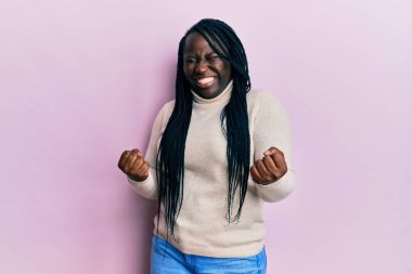 Saçları örgülü genç siyahi bir kadın, mutlu ve heyecanlı bir şekilde kolları havada, gülümseyen ve başarı için bağıran kazanan bir jest yapıyor. kutlama konsepti. 