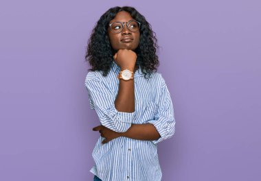 Güzel Afrikalı genç bir kadın, gündelik kıyafetler giyer ve elleri çenesinde gözlüklerle soru sorar, düşünceli bir ifade takınır. Gülümseyen ve düşünceli bir yüz. şüphe kavramı. 