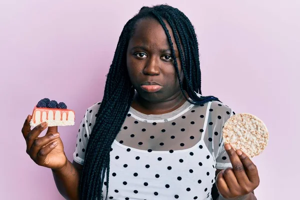 Young black woman with braids eating cake slice and rice crackers depressed and worry for distress, crying angry and afraid. sad expression.