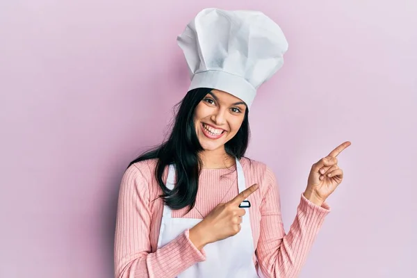 若いですヒスパニック女性身に着けていますパン屋の制服とコック帽子笑顔とカメラを見てポインティングで二本の手と指で側面 — ストック写真