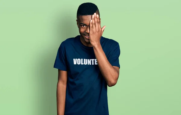アフリカ系アメリカ人の若者がボランティアのTシャツを着て 片手で片目を覆い 自信に満ちた笑顔と驚きの感情 — ストック写真
