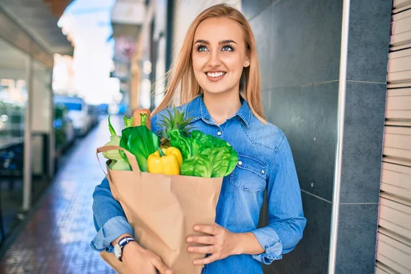 年轻的金发姑娘高兴地微笑着 手里拿着食品杂货纸袋站在城里 — 图库照片