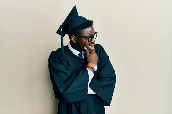 英俊的黑人男子戴着毕业典礼帽 身穿礼袍 满脑子思量着一个问题 手托着下巴忧心忡忡 — 图库照片