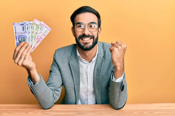 Joven Hispano Sosteniendo Billetes Rupias Indias Sentado Mesa Gritando Orgulloso —  Fotos de Stock