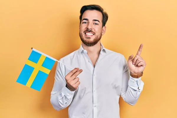 英俊的高加索人 留着胡子 举着瑞典国旗 高兴地微笑着 手指手画脚地指向旁边 — 图库照片