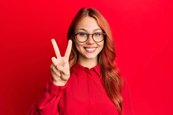 年轻美丽的红头发女人 身穿休闲装 戴着眼镜 穿着红色背景的衣服 带着自信和快乐的笑容 用手指指了指二号 — 图库照片