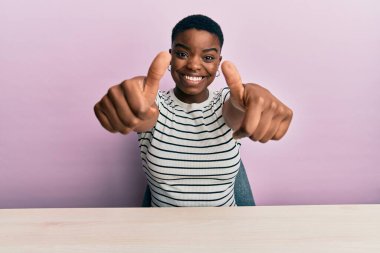 Genç Afro-Amerikalı kadın, gündelik giysiler içinde masaya oturmuş olumlu bir jest yapmayı onaylıyor, gülümseyerek ve başarı için seviniyor. kazanan jest. 