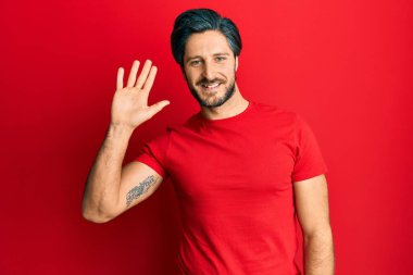 Latin asıllı genç adam, günlük kırmızı tişört giyiyor. Merhaba diyor, gülümsüyor ve gülümsüyor. Hoş geldin hareketi yapıyor. 