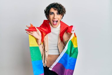Young man wearing woman make up wrapped in rainbow lgbtq flag celebrating crazy and amazed for success with open eyes screaming excited. 