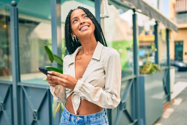 Genç Afrikalı Amerikalı kadın şehirde akıllı telefon kullanarak mutlu bir şekilde gülümsüyor..
