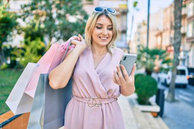 Genç sarışın kadın gülümsüyor alışveriş torbaları tutuyor ve şehirde akıllı telefon kullanıyor..