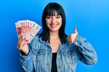 Young hispanic woman holding icelandic krona banknotes smiling happy and positive, thumb up doing excellent and approval sign 