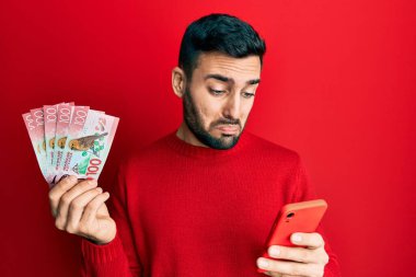 Genç İspanyol adam elinde yeni Zelanda dolarları tutan akıllı telefon kullanıyor. Depresyonda ve endişe içinde, sinirli ve korkmuş. hüzünlü ifade. 