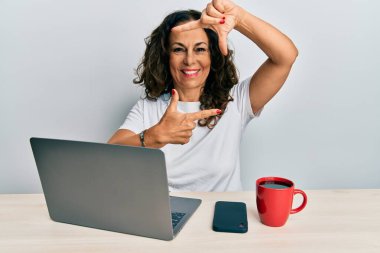 Ofiste çalışan orta yaşlı güzel bir kadın bilgisayarda dizüstü bilgisayar kullanıyor gülümsüyor ve mutlu yüzlü eller ve parmaklarla çerçeve yapıyor. yaratıcılık ve fotoğrafçılık kavramı. 