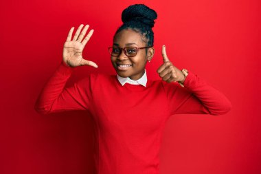 Genç Afro-Amerikalı kadın günlük kıyafetler ve gözlük takıyor. Altıncı parmaklarını gösterip işaret ederken kendinden emin ve mutlu bir şekilde gülümsüyor.. 