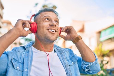 Genç beyaz adam şehirde kulaklık takarak mutlu bir şekilde gülümsüyor..