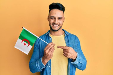 Cezayir bayrağı tutan genç Arap adam mutlu bir şekilde el ve parmağıyla işaret ediyor. 