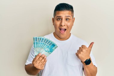 Elinde 100 Brezilyalı banknot tutan genç latin adam başparmağını yana kaldırmış, ağzı açık gülümsüyor. 