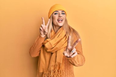 Genç beyaz kadın yün kazak ve şapkalı gülümseyen parmaklarını zafer işareti yaparken gösteren kameraya bakıyor. İki numara.. 