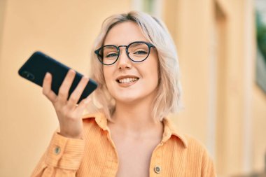 Genç sarışın kız gülümseyerek şehirde akıllı telefon kullanarak sesli mesaj gönderiyor..