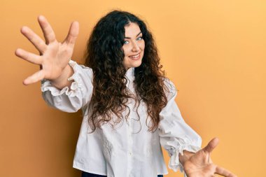 Kıvırcık saçlı, sıradan kıyafetler giyen genç esmer kadın sarılmak için kollarını açıp kameraya bakıp gülümsüyor. Mutluluğu kucaklayan neşeli bir ifade.. 