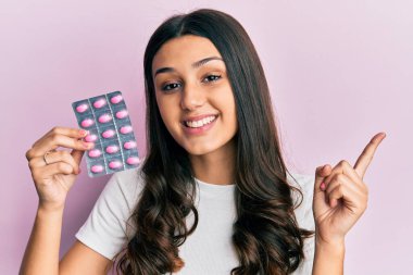 Genç İspanyol kadın elinde haplarla gülümsüyor mutlu bir şekilde elini ve parmağını yana doğru gösteriyor. 