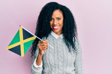 Orta yaşlı Afro-Amerikalı kadın elinde Jamaika bayrağı tutarken pozitif ve mutlu görünüyor ve kendine güvenen bir gülümsemeyle dişlerini gösteriyor. 