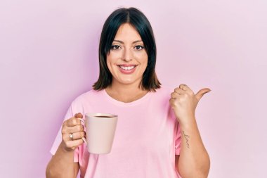 Genç İspanyol kadın bir fincan kahve içiyor başparmağıyla yan tarafa bakıyor mutlu bir şekilde gülümsüyor ve ağzı açık. 