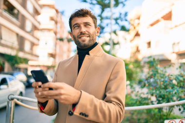 Zarif bir ceket giyen yakışıklı bir iş adamı akıllı telefon kullanarak dışarıda mutlu bir şekilde gülümsüyor.