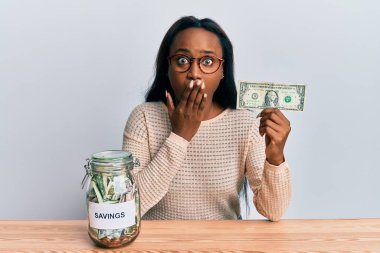 Elinde dolar ve kavanoz tutan genç Afrikalı bir kadın. Birikimleri ağzıyla kapatıyor. Şok olmuş ve hata yapmaktan korkuyor. şaşırmış ifade 