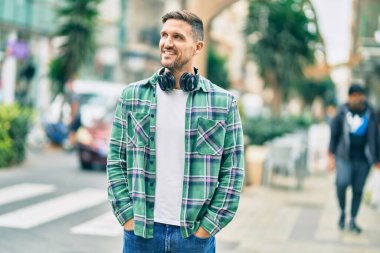 Genç beyaz adam şehirde kulaklık takarak mutlu bir şekilde gülümsüyor..