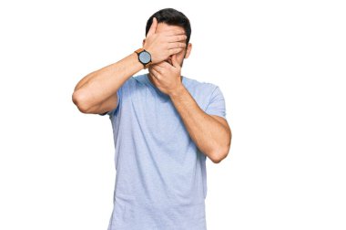 Gündelik kıyafetler giyen genç İspanyol adam gözlerini ve ağzını elleriyle kapatıyor, şaşırmış ve şok olmuş. duygu gizleme 