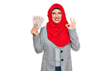 İslami tesettürlü güzel İspanyol kadın elinde 5 kilo banknot tutuyor. Parmaklarıyla imza atıyor, gülümseyerek mükemmel bir sembol oluşturuyor. 