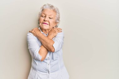 Kıdemli, gri saçlı, günlük kıyafetler giyen, kendine sarılan mutlu ve pozitif, kendinden emin gülümseyen bir kadın. Sevgi ve özgüven. 