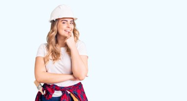 Güvenlik kaskı takan beyaz bir kadın kameraya kollarını kavuşturup gülümseyerek ve çenesini kaldırarak bakıyor. Olumlu düşünmek. 