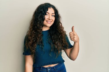 Kıvırcık saçlı, günlük kıyafetler giyen genç esmer kadın mutlu bir el hareketi yapıyor. Kameraya bakıp başarıyı gösteren ifade. 