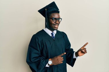 Mezuniyet şapkası ve tören cübbesi takan yakışıklı siyahi adam gülümsüyor ve kameraya bakıyor iki eliyle ve parmaklarıyla kenarı işaret ediyor.. 