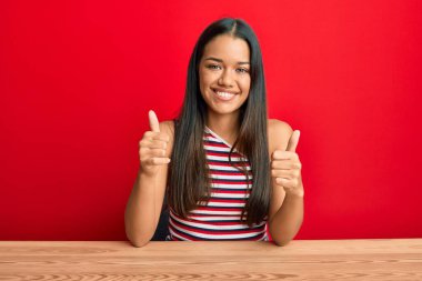 Güzel İspanyol kadın gündelik giysiler giyip masanın üzerinde oturuyor. Başparmağıyla gülümsüyor ve mutlu bir şekilde olumlu jest yapıyor. Neşeli ifade ve kazanan jest. 