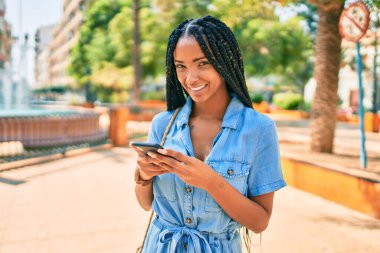 Genç Afrikalı Amerikalı kadın şehirde akıllı telefon kullanarak mutlu bir şekilde gülümsüyor..
