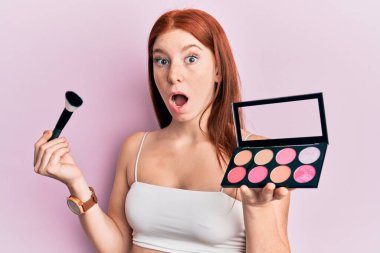 Makyaj fırçası ve allık paleti tutan genç kızıl saçlı kız şaşkınlık ve şaşkınlık dolu bir ifade, korku ve heyecanlı bir yüz ifadesiyle dehşete düşmüş ve şok olmuştu.. 