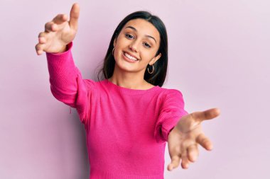 Gündelik giysiler giymiş genç İspanyol kadın kameraya bakıp sarılmak için kollarını açmış gülümsüyor. Mutluluğu kucaklayan neşeli bir ifade.. 