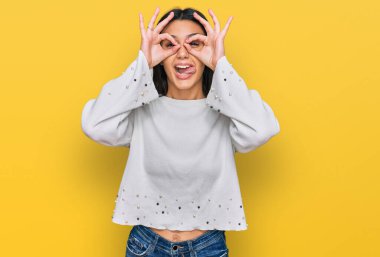 Günlük kıyafetler giyen genç İspanyol kız dürbün gibi jest yapıyor, gözleri parmaklarından bakıyor. çılgın ifade. 