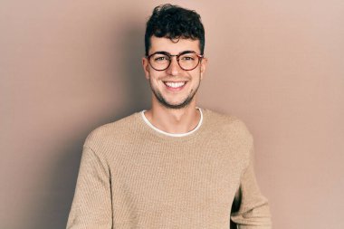 Gündelik kıyafetler ve gözlük takan genç İspanyol adam güler yüzlü ve kolları açık, dostça karşılama, olumlu ve kendinden emin selamlar. 